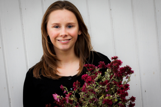 White woman with light brown hair holding a bouquet. She is smiling and wearing a black blouse.