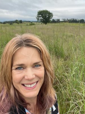 White woman with blonde hair has pink dyed ends. She is smiling in a green field with one tree.