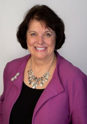 Smiling white woman with short brown hair. She is wearing a pink blazer and lovely silver necklace.