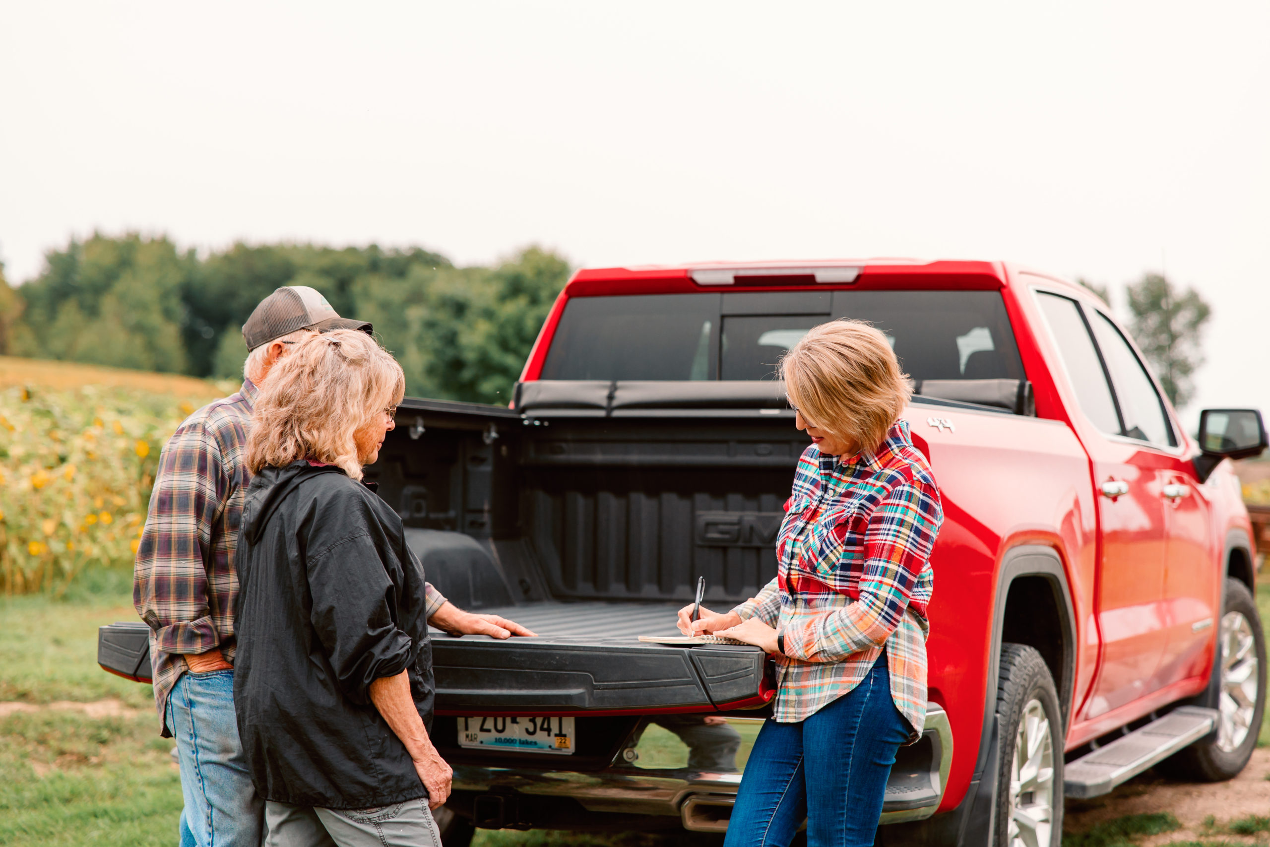 A picture of a woman signing a document on the back of a truck bed. The truck is red with Minnesota license plates. The woman signing the paper has blonde hair and is wearing a plaid shirt. The other woman has blonde hair and is wearing a black pull-over. The man is wearing a hat and a plaid shirt. The three people are near a farm with trees and a field in the background.