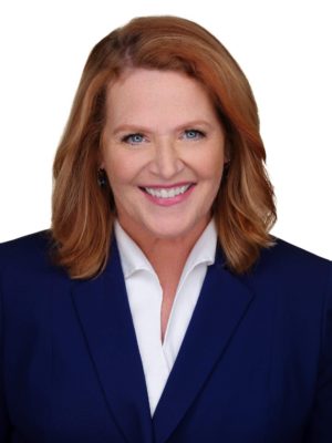 A picture of Heidi Heitkamp, she has short red hair and blue eyes. She is smiling and wearing a dark blazer with a white blouse.