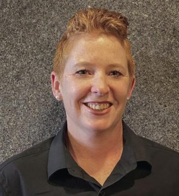 A picture of a woman with short red hair smiling against a textured, grey background. She is wearing a dark polo shirt.