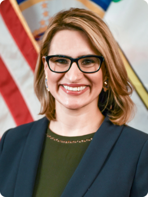 A picture of a woman with short blonde hair wearing glasses and smiling. She is wearing a green shirt with a blue blazer in front of an American flag.
