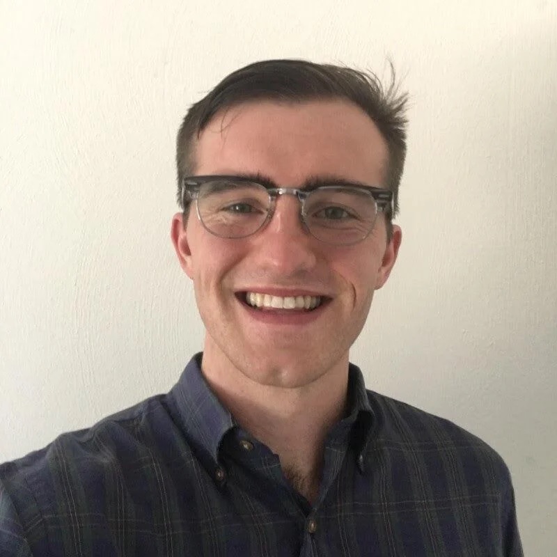 A picture of a white man smiling wearing glasses and a dark buttoned up shirt. He has light brown hair and stands in front of a plain, white wall.