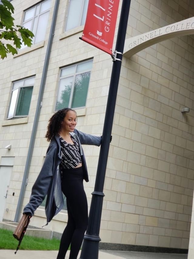 A picture of a tall woman with dark, curly hair wearing a jacket with a blouse and black jeans. She is smiling and hanging with one arm from a pole in front of a brick building.