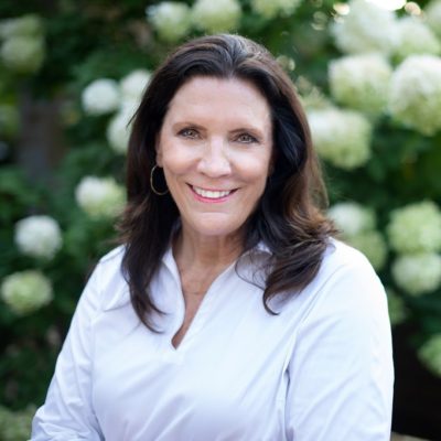 A picture of Minnesota State Senator, Julie Rosen. She has dark, mid-length hair. She is wearing and white blouse and is smiling in front of a flower bush.