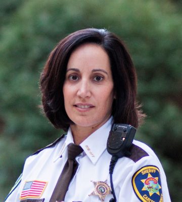 A picture of a woman smiling with short, dark brown hair. She is wearing a sheriff's uniform and is in front of green scenery.