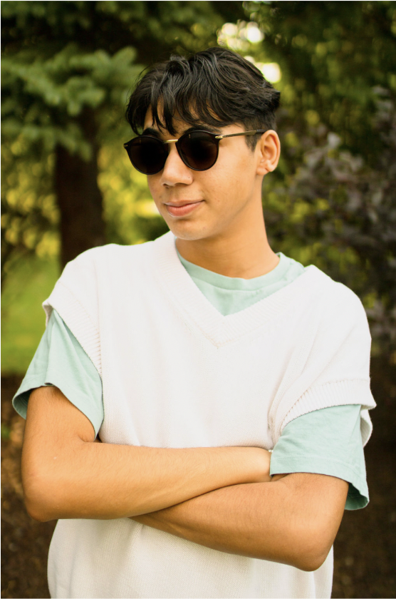 A picture of a boy wearing a light green t-shirt and white sweater vest. He is wearing sunglasses and has dark, short hair. He stands in front of trees.
