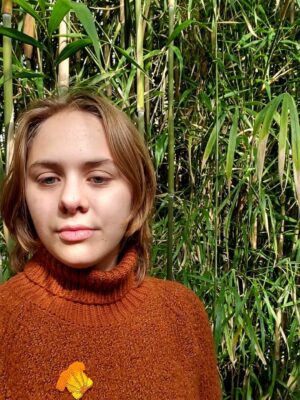 A picture of a girl with short blonde hair wearing a burnt orange turtleneck sweater in front of green scenery.