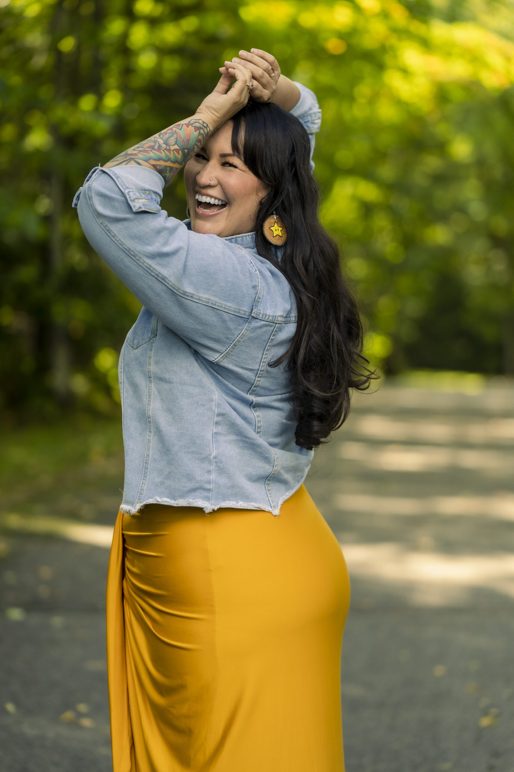 A tall woman stands sideways to the camera with her arms above her head. She is wearing a demin jacket and yellow skirt. She has long dark hair and is smiling.