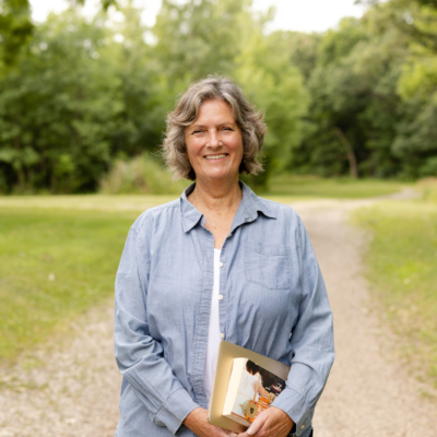 A picture of a woman wearing a jean shirt with a white top and black jeans. She is holding a book and smiling in front of a greenery background.