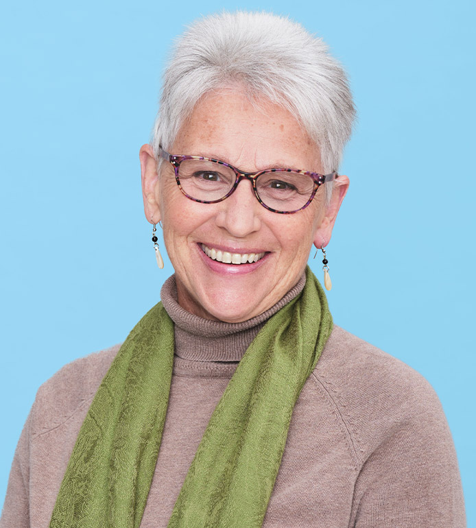 Suzanne has short grey hair and black framed glasses. She wears a green scarf over a grey turtle neck sweater.
